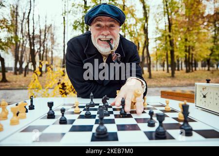 Ritratto dell'uomo anziano che gioca a scacchi nel parco di giorno in autunno. Look vincente. Concetto di attività di svago, vecchia generazione Foto Stock