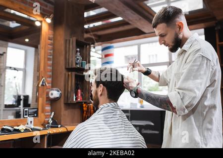 Barbiere tatuato pettinare i capelli del cliente e tenere forbici in barbershop, immagine stock Foto Stock