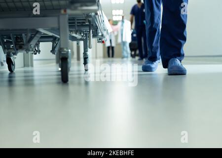 Sezione bassa delle gambe dei lavoratori ospedalieri che camminano e letto d'ospedale in corridoio, con spazio copia Foto Stock