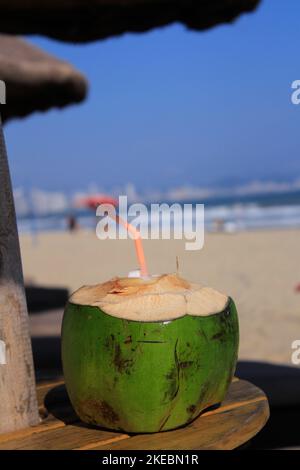 Un'acqua di cocco sul tavolo da spiaggia sotto l'ombrellone con una scena da spiaggia Foto Stock