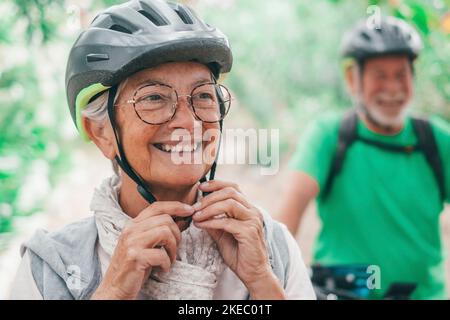 Ritratto di una vecchia donna sorridente e godendo la natura all'aperto in bicicletta con suo marito ridendo. Headshot di femmina matura con gli occhiali che si sentono sani. Indossare il casco per andare in bicicletta. Foto Stock