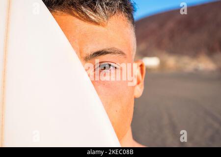 primo piano e ritratto di un giovane e bell'uomo con una tavola da surf che copre la metà del suo volto - stile di vita attivo adolescente Foto Stock