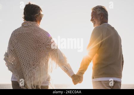 coppia di due anziani felici e attivi divertirsi e godersi insieme l'estate in spiaggia a piedi tenendo le mani con il tramonto sullo sfondo Foto Stock
