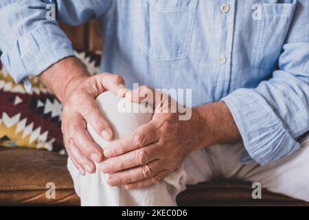 Primo piano dell'anziano che affronta il problema del ginocchio, seduto sul divano che tiene il ginocchio a casa. Uomo anziano che soffre di grave dolore al ginocchio seduto a casa Foto Stock