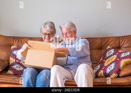 Vecchia coppia disimballare la scatola di consegna a casa. Coppia anziana felice che guarda la scatola di cartone mentre si siede sul divano nel soggiorno a casa. La coppia anziana che controlla fuori ha trasportato il roba nel paese. Foto Stock