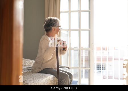 Donna anziana premurosa che si appoggia sul bastone mentre si siede sul letto. Donna anziana solitaria seduta in camera da letto a casa. Vecchia donna con i capelli grigi appoggiati sul bastone che cammina seduto sul letto a casa Foto Stock