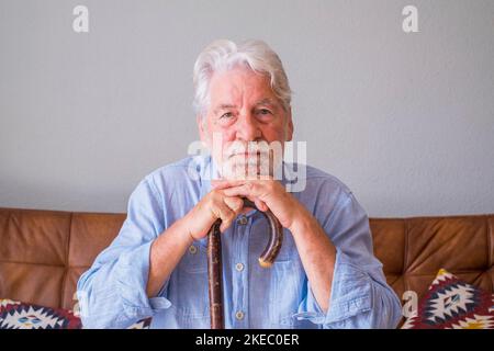 Ritratto dell'uomo anziano che si appoggia sul bastone da passeggio mentre si siede sul divano. Solitario uomo anziano seduto nel soggiorno di casa. Vecchio con i capelli grigi che si appoggiano sul bastone che cammina seduto sul divano a casa Foto Stock