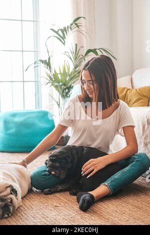 Ritratto di bella donna in occhiali che si divertono con i suoi animali domestici pugs cani seduti sul pavimento nel soggiorno della sua casa. Donna allegra trascorrere il tempo libero con i suoi due cani carino a casa Foto Stock