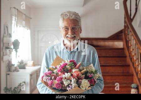 Ritratto di un vecchio felice e carino che tiene fiori da dare a sua moglie o fidanzate. Senior guardando la macchina fotografica a casa divertirsi con regali o regali per San Valentino Foto Stock