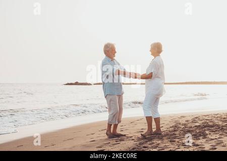 Coppia di persone anziane che ballano insieme e che si divertono sulla sabbia in spiaggia godendosi e vivendo il momento. Ritratto degli anziani innamorati che si guardano gli altri divertendosi. Foto Stock