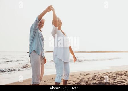 Coppia di persone anziane che ballano insieme e che si divertono sulla sabbia in spiaggia godendosi e vivendo il momento. Ritratto degli anziani innamorati che si guardano gli altri divertendosi. Foto Stock