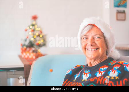 Ritratto di donna anziana sorridente in pensione in cappello di santa e abiti caldi che celebrano il natale a casa. Santa vecchia donna sorridente mentre guardava la fotocamera. Donna anziana che ama le vacanze di natale Foto Stock