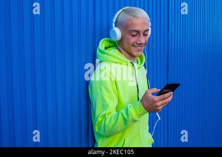 primo piano e ritratto di un giovane adolescente che ascolta musica e si diverte con il suo telefono e indossando le cuffie - parete di sfondo blu - allegra e felice millennial ascoltare musica e sorrisi Foto Stock