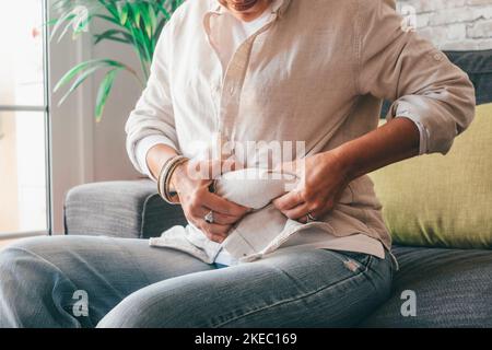 Parte centrale delle mani della donna che tiene il suo grasso della pancia seduto sul divano a casa. Donna caucasica afferrare troppo grasso sul suo addome. Giovane femmina che stringe la sua vita obesa grassa Foto Stock