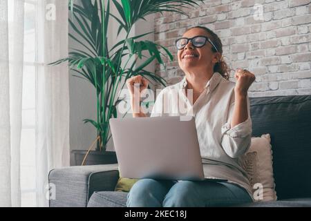 Donna seduto sul divano mettere il pc in giro pugni clenched grida di gioia, mentre leggere grandi notizie sul notebook. Il giocatore celebra la vittoria della scommessa dell'asta in linea. Ha ottenuto l'offerta incredibile emozioni sincere del concetto di felicità Foto Stock