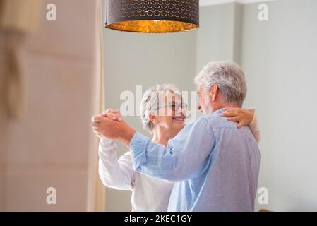 Coppia anziana amorevole romantica che tiene le mani godendo ballare insieme nel soggiorno di casa, coppia anziana felice che festeggia facendo la danza a casa. Vecchio marito e moglie che si divertono in un appartamento moderno Foto Stock