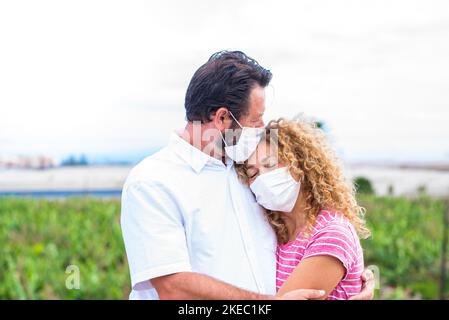 coppia di persone abbracciato insieme e l'uomo che dà un bacio a sua moglie - due adulti che indossano una maschera medica e chirurgica su di loro faccia per prevenire il covid-19 o qualsiasi tipo di malattia o influenza - nel concetto di amore Foto Stock