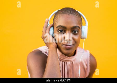 Ritratto di donna con pelle scura che ascolta musica sulle cuffie su sfondo giallo. Sorridente ragazza afroamericana godendo la sua canzone preferita utilizzando apparecchiature audio via cavo. Tecnologia e intrattenimento Foto Stock