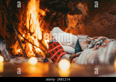 Gambe di marito e moglie a sezione bassa coperte di calze e coperta calda rilassante di fronte al camino in fiamme durante le vacanze invernali di natale. Paio di piedi in calze di lana vicino al camino di Natale Foto Stock