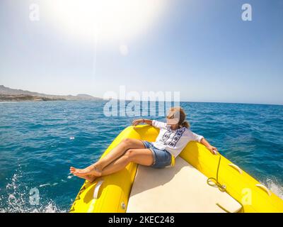 una giovane donna sdraiata su una barca che viaggia alla scoperta del mare godendosi e rilassandosi con una giornata di sole - guardando la costa dal gommone Foto Stock