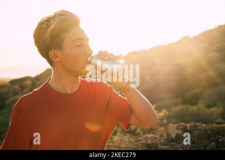 ritratto e primo piano di un giovane o di un adolescente che beve acqua e che sbatte con l'acqua la sera con il tramonto sullo sfondo Foto Stock