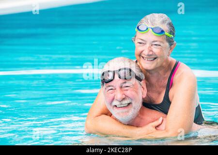 coppia di due anziani nella piscina per divertirsi insieme sorridendo e nuotando - donna matura abbracciando dal indietro suo marito - amore e affetto di persone attive all'aperto in piscina Foto Stock