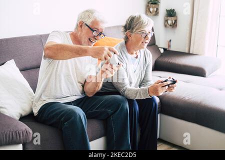 coppia di persone anziane e mature che guardano la tv e usano i controller che giocano insieme a casa i videogiochi sul divano - stile di vita chiuso al chiuso Foto Stock