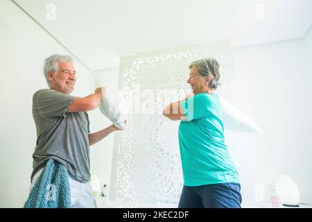 Maturo pensionato felice coppia invecchiata divertirsi a casa con cuscini in camera da letto - stile di vita gioioso per molto tempo persone sposate - allegro anziano uomo e donna caucasica - lockdown divertirsi in quarantena Foto Stock