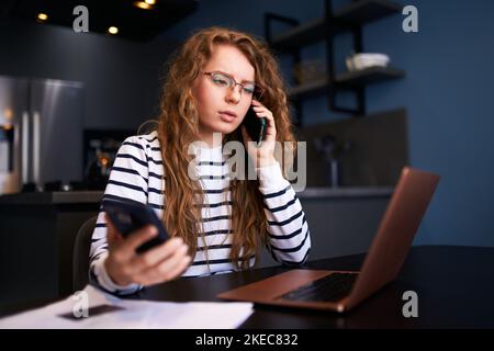 Donna bionda riccia in occhiali che lavora da casa con più dispositivi elettronici Internet. Freelancer donna d'affari tiene tablet, parlando sul cellulare Foto Stock