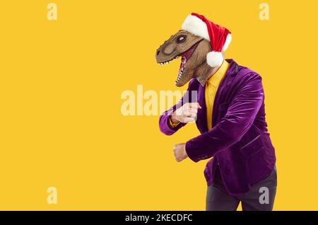 Uomo divertente in maschera di dinosauro, cappello di Natale e abito da festa ballare su sfondo spazio copia Foto Stock