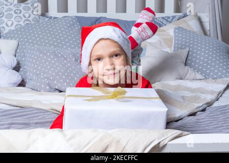 Una ragazza in pigiama rossa e un cappello di Babbo Natale tiene una grande scatola regalo, sdraiata sul letto nella stanza dei bambini. Regali di Natale e Capodanno per i bambini. Famiglia Chris Foto Stock