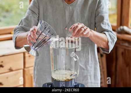 Passaggi per la preparazione del latte di mandorla, una tazza dosatrice d'acqua e una ciotola di vetro di sciroppo d'acero sono messi da una donna in un frullatore riempito con mandorle sbiancate Foto Stock