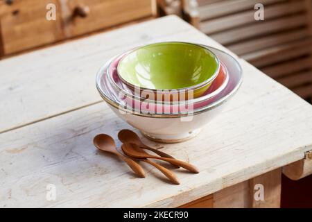 Ciotole di diversi colori e dimensioni sono impilate l'una sull'altra su un tavolo di legno grezzo, accanto a quattro cucchiai di legno Foto Stock