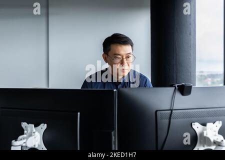 Un serio giovane asiatico lavora al computer in un ufficio moderno. Si siede concentrato al tavolo vicino alla finestra. Foto Stock
