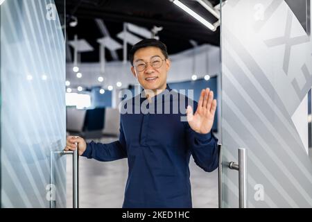 Un giovane e bello architetto asiatico, designer, ingegnere si trova alla porta di un ufficio moderno, saluta, sorride alla fotocamera. Foto Stock