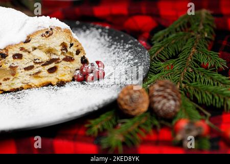 Un pezzo tagliato di Natale tedesco stollen su un piatto nero decorato con una tovaglia rossa e nera, coni di pino, bacche, rami di albero di Natale. Foto Stock