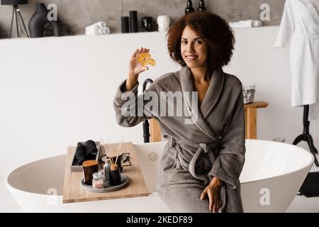 Donna africana che tiene le zone dell'occhio dell'oro per lisciare le rughe, rimuovere il gonfiamento o le borse sotto gli occhi. Ragazza in accappatoio con occhielli in idrogel d'oro w Foto Stock