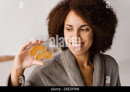 Donna africana che tiene le zone dell'occhio dell'oro per lisciare le rughe, rimuovere il gonfiamento o le borse sotto gli occhi. Ragazza in accappatoio con occhielli in idrogel d'oro w Foto Stock