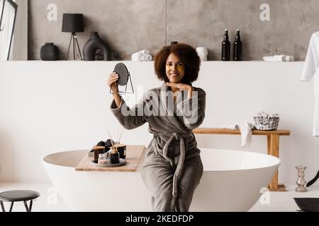 Ragazza africana in accappatoio con occhielli dorati sta guardando lo specchio in bagno. Mattina bellezza routine di donna afro-americana Foto Stock