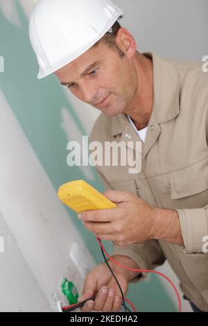 elettricista che controlla la tensione in una presa elettrica a muro parzialmente installata Foto Stock