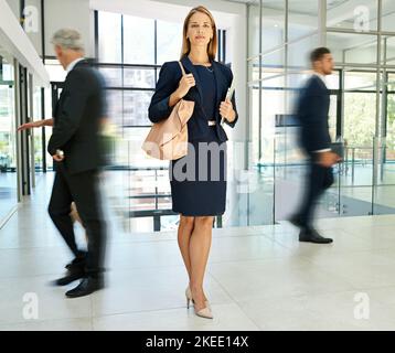 Rimanere fermi mentre tutti gli altri sono occupati. Scatto a tutta lunghezza di una giovane e attraente donna d'affari in piedi in un ufficio moderno mentre i suoi colleghi Foto Stock