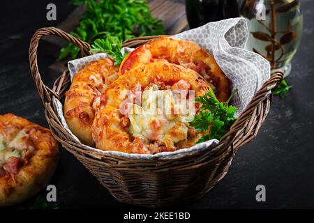 Mini pizza per bambini, piccole torte aperte con prosciutto, salsa di pomodoro, mozzarella e formaggio in cestino su fondo scuro. Foto Stock