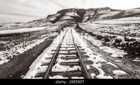 Treno binari ferroviari in montagne rurali coperte di neve d'inverno closeup sopra una seppia scenografica bianco e nero contrastante paesaggio di viaggio. Foto Stock