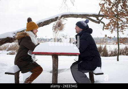 felici amanti uomo e donna, lui e lei in abiti caldi d'inverno, sedersi a un tavolo coperto di neve in un parco d'inverno. passeggiate invernali. godere il momento in un Foto Stock