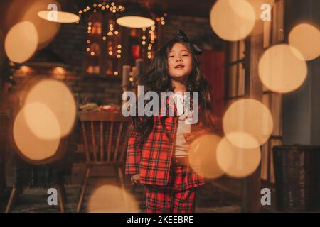 Attiva ragazza bambino giappone con elegante modello di capelli brunette lunga posa a casa in bokeh ghirlands noel. Asian piccolo fashionista in rosso plaid pajama danza Foto Stock