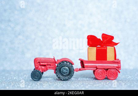Biglietto d'auguri: Il trattore giocattolo rosso trasporta una grande scatola regalo avvolta su uno sfondo scintillante che imita la neve. Foto Stock