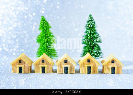 Fila di case in miniatura poste di fronte a due alberi di Natale giocattolo su sfondo scintillante. Foto Stock