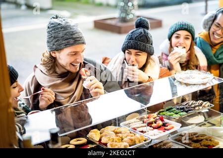 Amici felici che si divertono a mangiare al mercato di Natale nel periodo invernale - Urban Holiday Concept con i giovani che si aggrappano insieme da negozio di caramelle indossare Foto Stock