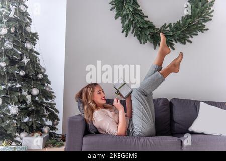Donna millenaria con rossetto rosso in stile pin-up, tenendo un dono in mano, gambe sollevate su un divano grigio vicino all'albero di Natale, corona. Nuovo pazzo Foto Stock