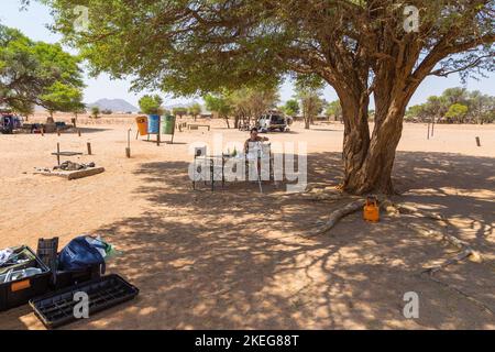 Sesriem Campsite, Sossusvlei, Namibia - 30 settembre 2018: Una ragazza seduta sotto un albero di acacia nel campeggio. Auto con attrezzature parcheggiate nelle vicinanze. Foto Stock